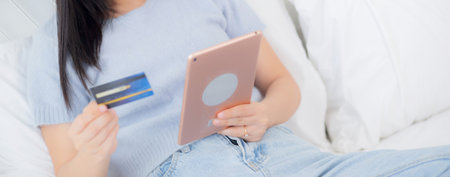 Young asian woman sitting on bed using digital tablet shopping online with credit card in the...