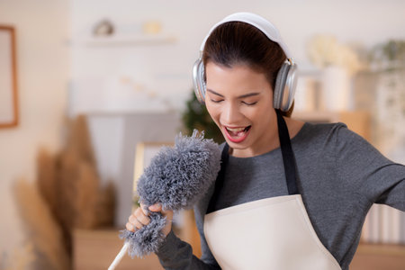Young asian woman dusting while listening to music with headphones in a bright, organized living room, maid doing housework enjoying music and sing while dusting at home, lifestyle concept.の写真素材