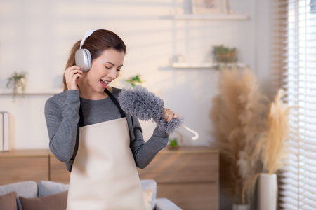 Young asian woman dusting while listening to music with headphones in a bright, organized living...