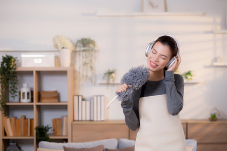 Young asian woman dusting while listening to music with headphones in a bright, organized living...