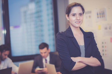 Professional businesswoman standing confident in modern office with colleagues meeting background,...