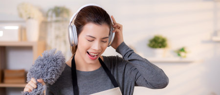Young asian woman dusting while listening to music with headphones in a bright, organized living room, maid doing housework enjoying music and sing while dusting at home, lifestyle concept.の写真素材
