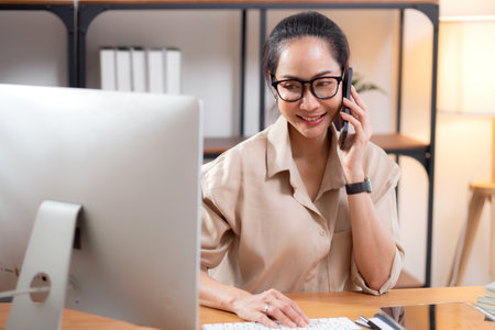 Young asian business woman using desktop computer while talking with smart phone in home office, businesswoman working with desktop computer while speaking with telephone, business or consulting.の写真素材