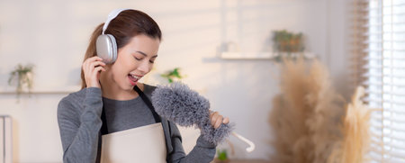 Young asian woman dusting while listening to music with headphones in a bright, organized living...