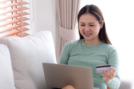 Young asian woman sitting on sofa using laptop computer shopping online with credit card buying to internet, female payment with business on couch, purchase and payment, business and lifestyles.の写真素材
