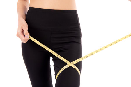 Closeup young asian woman in sportswear holding measuring tape leg for weight loss isolated white background, girl weightloss with workout and exercise while measurement with leg, sport and diet.の写真素材
