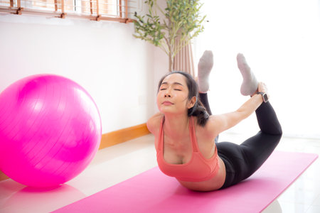 Young asian woman practicing Yoga with stretching muscle back on yoga mat in living room at home, woman exercise pilates with challenge and motivation, workout for wellness, sport and wellbeing.の写真素材