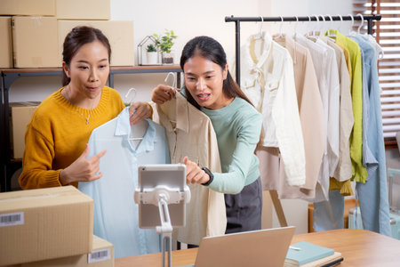 Two young asian woman live online clothing store while showing clothing with tablet on social at home, entrepreneur streaming for selling online clothing and presenting, online seller.の写真素材