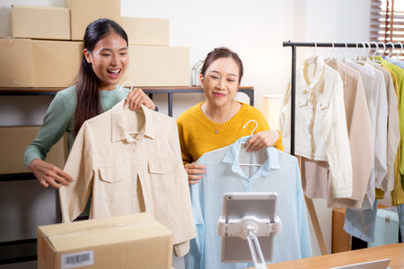 Two young asian woman live online clothing store while showing clothing with tablet on social at home, entrepreneur streaming for selling online clothing and presenting, online seller.の写真素材