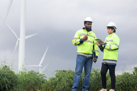 Two engineer check and examining wind turbines in wind farm, renewable energy and teamwork in a sustainable environment, inspection power electric with document and tablet, industry and eco.の写真素材