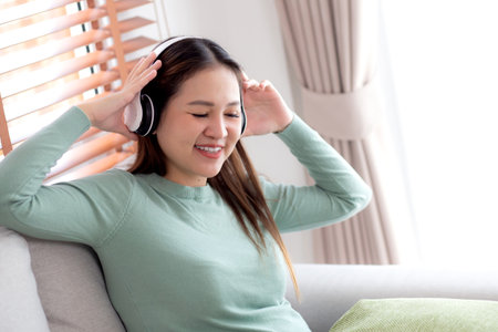 Happy young asian woman wearing headphones listening music while sitting on sofa with fun and enjoy in the living room at home, happiness female with entertainment, one person, lifestyle concept.の写真素材