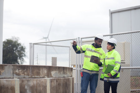 Two engineer check and examining wind turbines in wind farm, renewable energy and teamwork in a sustainable environment, inspection power electric with document and tablet, industry and eco.の写真素材