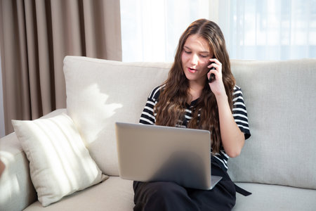Young caucasian businesswoman working on laptop computer sitting on sofa while talking on smart phone in living room at home office, business woman using laptop while talking on smartphone.の写真素材