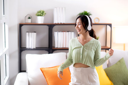 Happy young asian woman wearing headphones listening music while standing dancing with fun and enjoy in the living room at home, happiness female with entertainment, one person, lifestyle concept.の写真素材