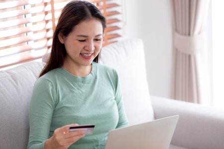 Young asian woman sitting on sofa using laptop computer shopping online with credit card buying to internet, female payment with business on couch, purchase and payment, business and lifestyles.の写真素材