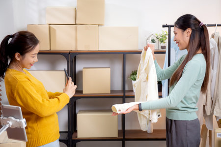 Two asian woman taking picture of clothes for online store, woman collaboration with online seller taking photo clothes to social media, businesswoman or entrepreneur selling product, small business.の写真素材