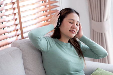 Happy young asian woman wearing headphones listening music while sitting on sofa with fun and enjoy in the living room at home, happiness female with entertainment, one person, lifestyle concept.の写真素材