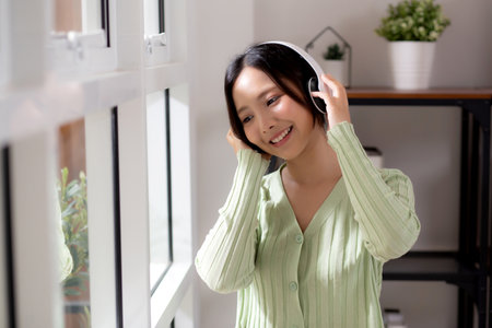 Portrait happy young woman wearing headphones listening music while standing with fun and enjoy in the living room at home, happiness female with entertainment, one person, lifestyle concept.の写真素材