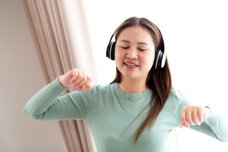 Happy young asian woman wearing headphones listening music while standing dancing with fun and enjoy in the living room at home, happiness female with entertainment, one person, lifestyle concept.の写真素材
