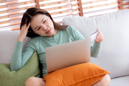 Young asian woman holding credit card or debit card with stress about loan and expense sitting on sofa in living room at home, woman worried and frustration about budget and finance, business concept.の写真素材
