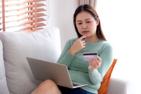 Young asian woman sitting on sofa using laptop computer shopping online with credit card buying to internet while thoughtful, female payment with business, purchase and payment, business and finance.の写真素材