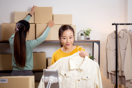 Two young asian woman live online clothing store while showing clothing with tablet and laptop on social at home, entrepreneur streaming for selling online clothing and presenting, online seller.の写真素材