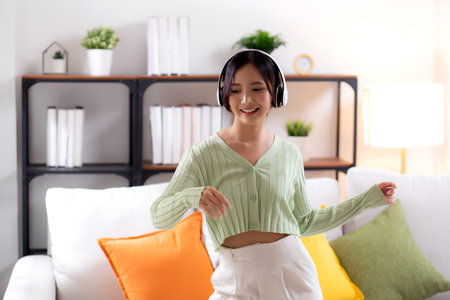 Happy young asian woman wearing headphones listening music while standing dancing with fun and enjoy in the living room at home, happiness female with entertainment, one person, lifestyle concept.の写真素材