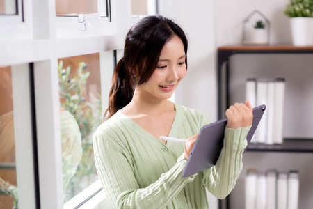 Young asian woman using tablet writing with inspiration and comfortable by window in living room at home office, cheerful woman standing working on digital tablet in workspace, business and lifestyle.の写真素材