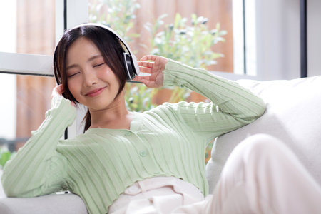 Happy young asian woman wearing headphones listening music while sitting on sofa with fun and enjoy in the living room at home, happiness female with entertainment, one person, lifestyle concept.の写真素材