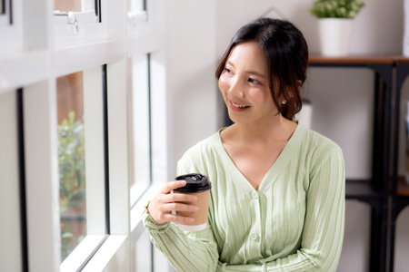 Portrait young asian woman relax and enjoyment with drinking coffee by the window in living room at home, woman smiling with happiness and comfortable drinking beverage at home, lifestyle and leisure.の写真素材
