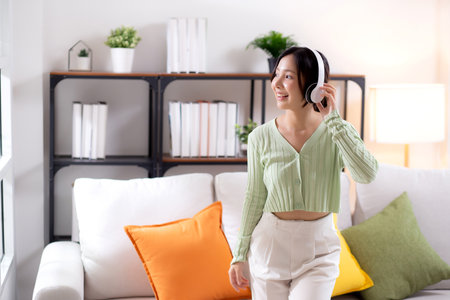 Happy young asian woman wearing headphones listening music while standing dancing with fun and enjoy in the living room at home, happiness female with entertainment, one person, lifestyle concept.の写真素材