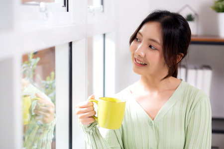 Portrait young asian woman relax and enjoyment with drinking coffee by the window in living room at home, woman smiling with happiness and comfortable drinking beverage at home, lifestyle and leisure.の写真素材