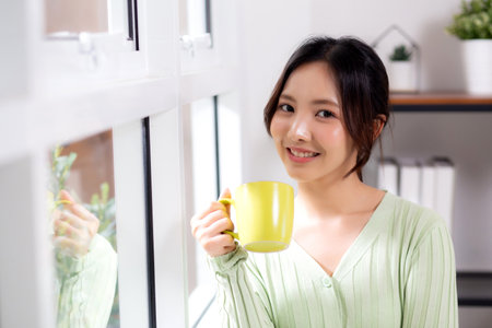 Portrait young asian woman relax and enjoyment with drinking coffee by the window in living room at home, woman smiling with happiness and comfortable drinking beverage at home, lifestyle and leisure.の写真素材