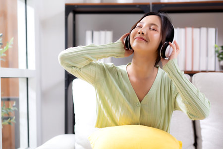 Happy young asian woman wearing headphones listening music while sitting on sofa with fun and enjoy in the living room at home, happiness female with entertainment, one person, lifestyle concept.の写真素材