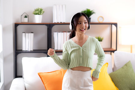 Happy young asian woman wearing headphones listening music while standing dancing with fun and enjoy in the living room at home, happiness female with entertainment, one person, lifestyle concept.の写真素材