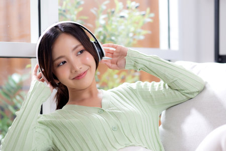 Happy young asian woman wearing headphones listening music while sitting on sofa with fun and enjoy in the living room at home, happiness female with entertainment, one person, lifestyle concept.の写真素材