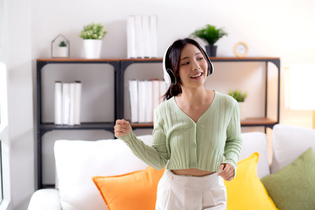 Happy young asian woman wearing headphones listening music while standing dancing with fun and enjoy in the living room at home, happiness female with entertainment, one person, lifestyle concept.の写真素材