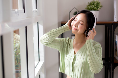 Portrait happy young woman wearing headphones listening music while standing with fun and enjoy in the living room at home, happiness female with entertainment, one person, lifestyle concept.の写真素材