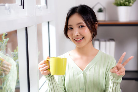Portrait young asian woman relax and enjoyment with drinking coffee by the window in living room at home, woman smiling with happiness and comfortable drinking beverage at home, lifestyle and leisure.の写真素材