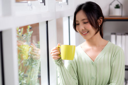 Portrait young asian woman relax and enjoyment with drinking coffee by the window in living room at home, woman smiling with happiness and comfortable drinking beverage at home, lifestyle and leisure.の写真素材