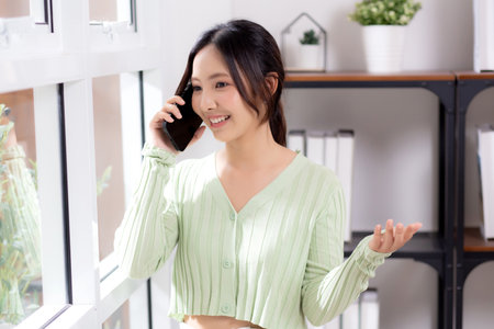 Young asian businesswoman telephone smiling and relax in living room at home office, cheerful woman standing talking with smart phone in home, expression and emotion, business and communication.の写真素材