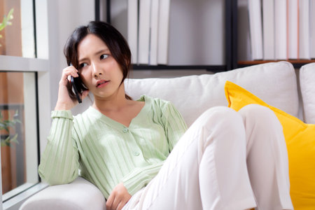 Asian young woman stress and worried sitting on sofa while speaking on smart phone in living room at home, woman unhappy and concerned talking on smartphone, lifestyle and communication.の写真素材