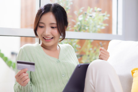 Young asian woman sitting on sofa using tablet computer shopping online with credit card buying to internet while cheerful, female payment with business, purchase and payment, business and finance.の写真素材