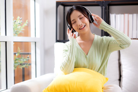 Happy young asian woman wearing headphones listening music while sitting on sofa with fun and enjoy in the living room at home, happiness female with entertainment, one person, lifestyle concept.の写真素材