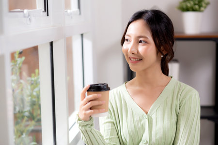 Portrait young asian woman relax and enjoyment with drinking coffee by the window in living room at home, woman smiling with happiness and comfortable drinking beverage at home, lifestyle and leisure.の写真素材