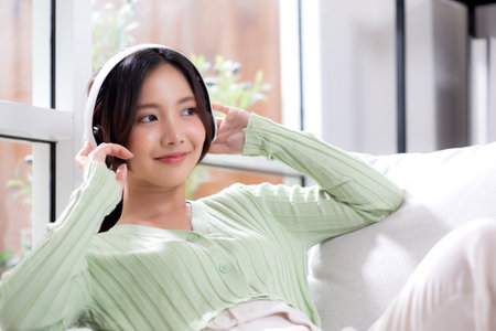 Happy young asian woman wearing headphones listening music while sitting on sofa with fun and enjoy in the living room at home, happiness female with entertainment, one person, lifestyle concept.の写真素材