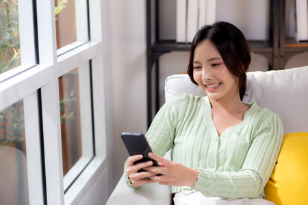 Young asian businesswoman looking smartphone smiling and relax sitting on sofa in living room at home, cheerful woman watching smart phone sitting on couch in home, lifestyle and communication.の写真素材