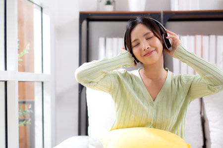 Happy young asian woman wearing headphones listening music while sitting on sofa with fun and enjoy in the living room at home, happiness female with entertainment, one person, lifestyle concept.の写真素材