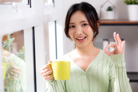 Portrait young asian woman relax and enjoyment with drinking coffee by the window in living room at home, woman smiling with happiness and comfortable drinking beverage at home, lifestyle and leisure.の写真素材