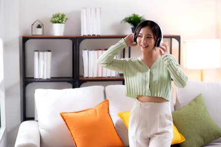 Happy young asian woman wearing headphones listening music while standing dancing with fun and enjoy in the living room at home, happiness female with entertainment, one person, lifestyle concept.の写真素材